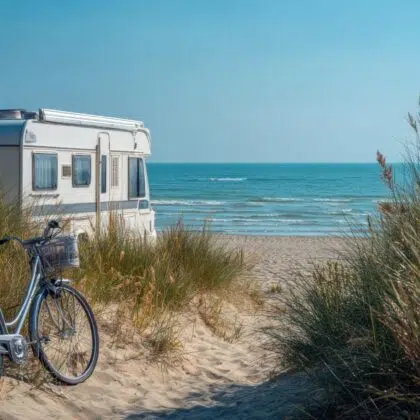 vendée en camping-car 6 White caravan, bicycle are parked on a sandy beach path, with beach grass and dunes, with the calm blue ocean stretching out under a clear sky in the background. Perfect for themes of summer vacation, outdoor adventure, travel, or a tranquil escape